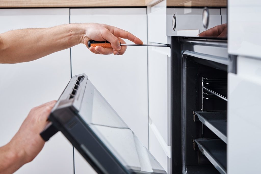A person using a screwdriver to repair a white built-in oven in a modern kitchen.