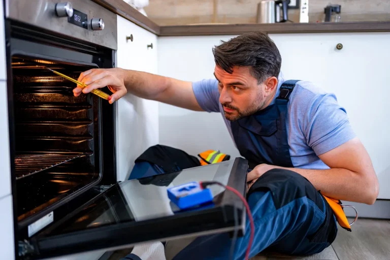 A technician in blue overalls sits on a kitchen floor, using a voltage tester and screwdriver to inspect the interior of an oven.