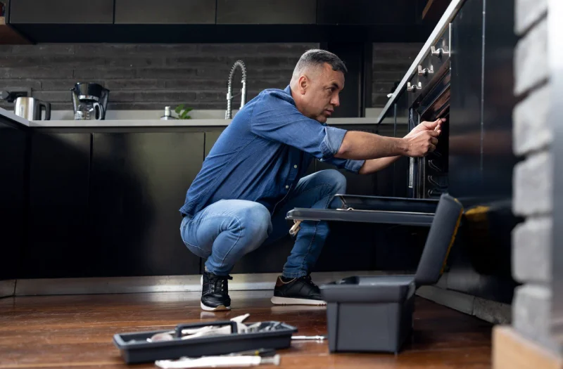 A technician crouches in a dark modern kitchen for appliance troubleshooting with tools scattered on the floor nearby.