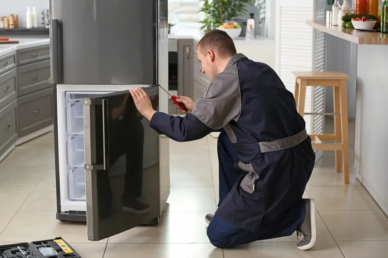 A local appliance repair expert in a uniform kneeling to repair a dark grey refrigerator in a kitchen.