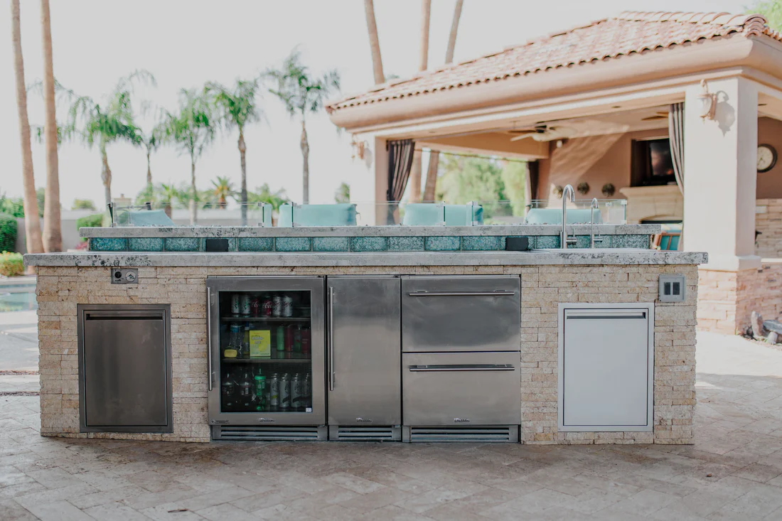 Modern outdoor kitchen island with a stone finish and stainless steel beverage fridge, set in a tropical backyard with palm trees and a poolside cabana.